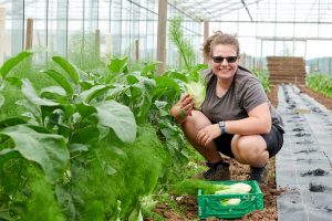 eine junge Frau hockt im Gewächshaus neben einem Beet und erntet Fenchel